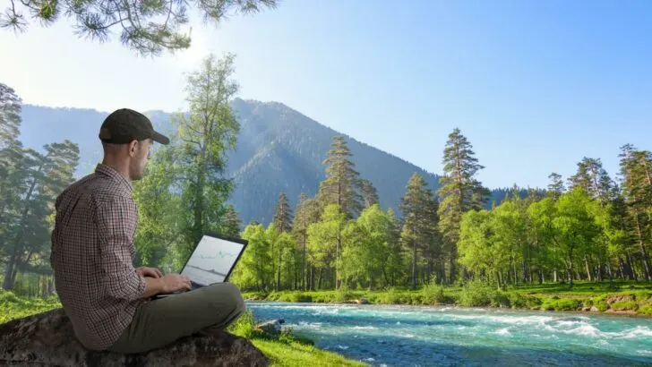A young man workamping on a computer beside a stream