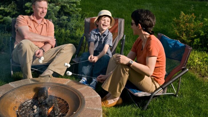A man, woman, and child sitting in camp chairs by a fire pit roasting marshmallows