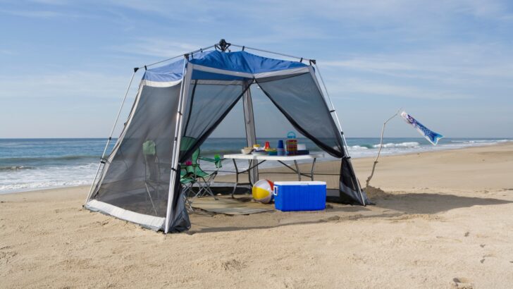 A screen tent for campers set up on the beach with table, cooler, toys, etc.