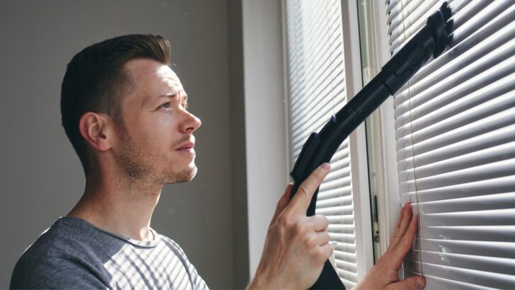 A man using a brush tool to vacuum window blinds