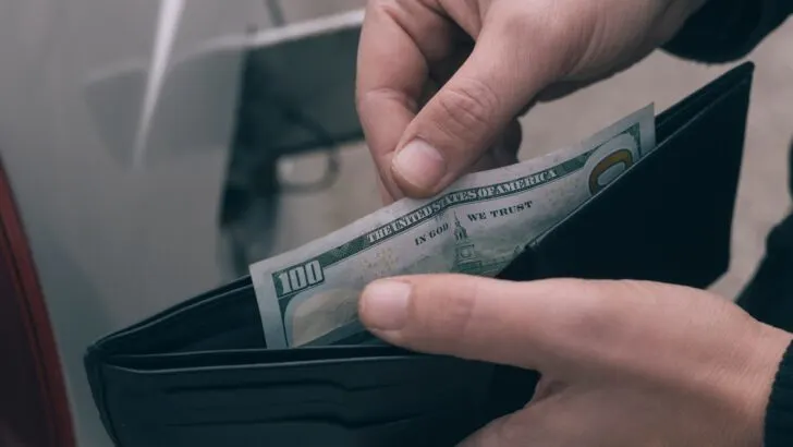 A man's hand retrieving a 100-dollar bill from a wallet at a fueling station